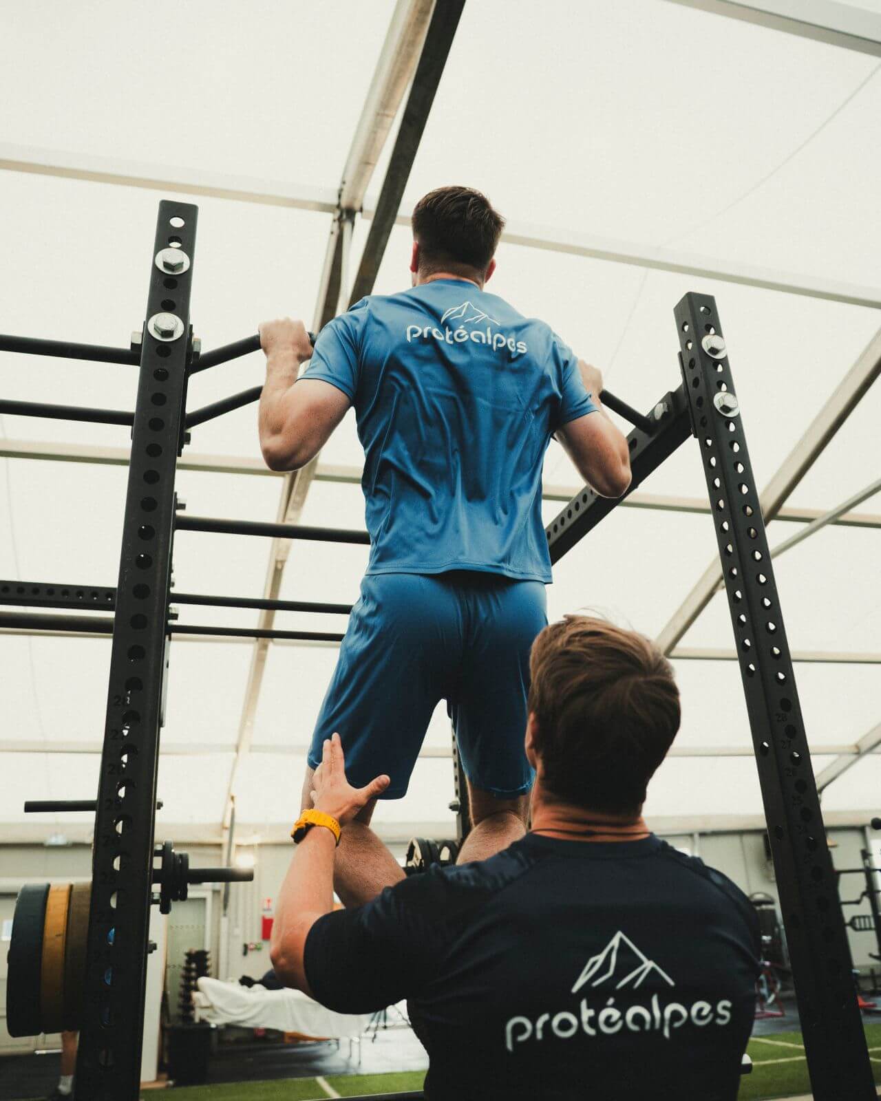 Two athletes, wearing Protéalpes jerseys, train at the high bar in a modern gym.