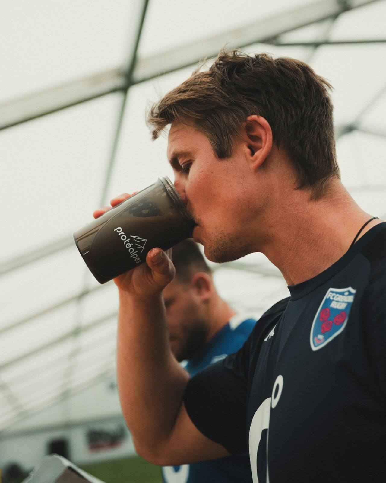 Athlete drinking a Protéalpes protein shake, sports nutrition without sweeteners or additives, under a metal structure.