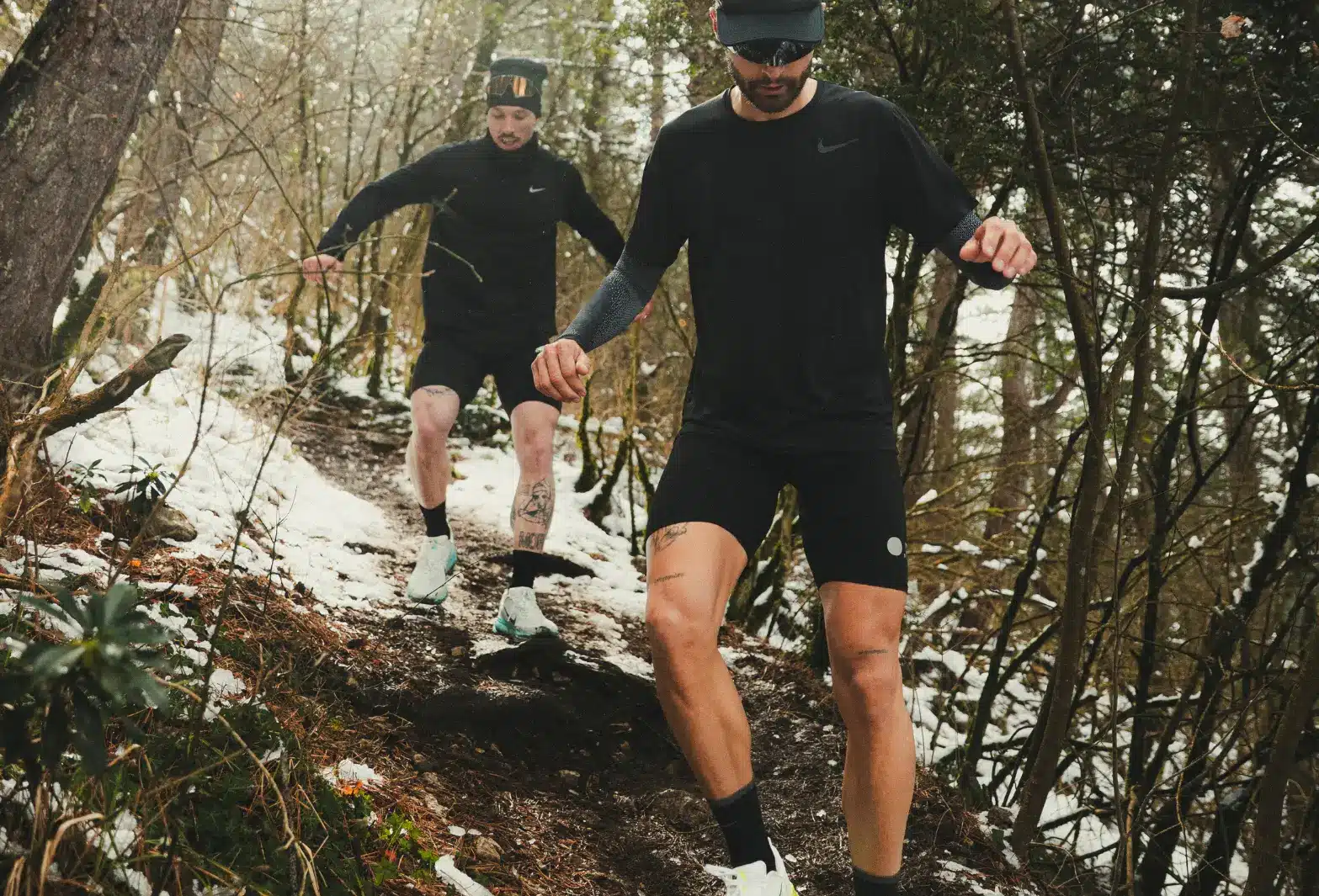 Deux coureurs en tenue noire descendent un sentier boisé enneigé, illustrant le lien entre nature et performance sportive.